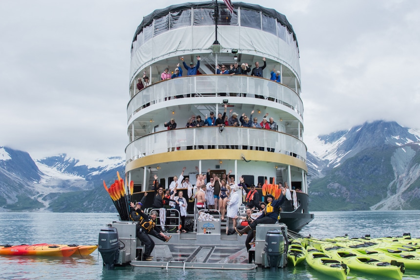 Guests and crew wave from the back of the 4 level boat, the Wilderness Adventurer, as it floats in Alaska surrounded by a mountain range. 