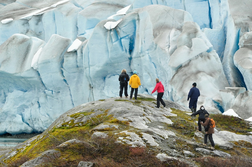In Alaska a group of hikers walk to a vista point with up-close views of a massive white and teal icy glacier. 