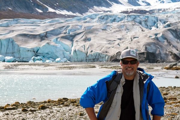 A small ship Alaska cruise specialist poses in front of glaciers wearing a blue jacket and grey hat with AdventureSmith Explorations logo