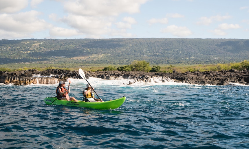 Two travelers paddle along the shore line of a Hawaiian Island in a neon green double kayak