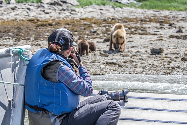 A woman in a plaid shirt, blue lifevest and black hat sits in a small ship Alaska cruise skiff photographing a brown bear.