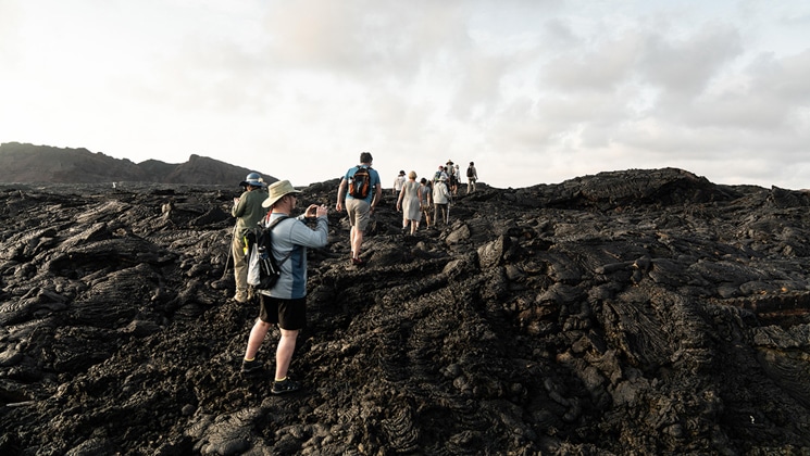 Small group of hikers walks over dark lava rock & takes pictures on a cloudy day during the Aqua Mare Galapagos Cruise.