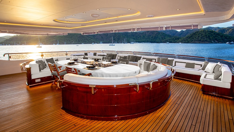 Aft deck with covered al fresco dining on Aqua Mare Galapagos superyacht, with teak deck, veneer oval seating bench & white cushions.