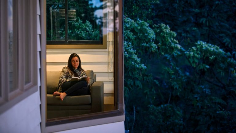 Woman sits on couch & reads beside large view windows overlooking cloud forest at Senda Monteverde eco-hotel in Costa Rica.