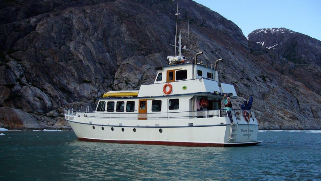 Exterior of Snow Goose Alaska yacht sitting in calm water beside rocky cliffs, with white walls, red hull & 3 decks.