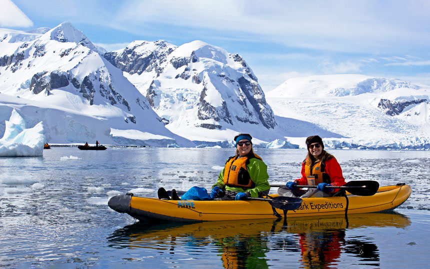 Floating in a yellow double kayak two guests wear green and red dry suits and pose in front of a snowy Antarctica mountain range.