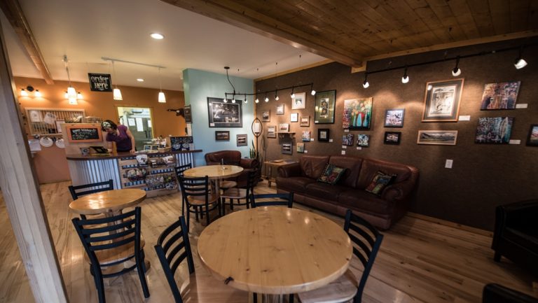 Tonglen Lake Denali lodge Artisan cafe with framed paintings hung, round wooden tables & service bar in the background.