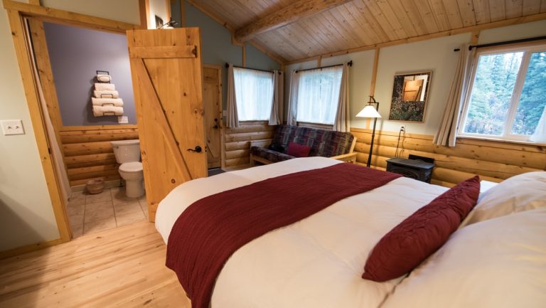 Tonglen Lake Denali lodge cabin interior with futon, king bed, wood accents, bright windows, lamp & small cast iron stove.