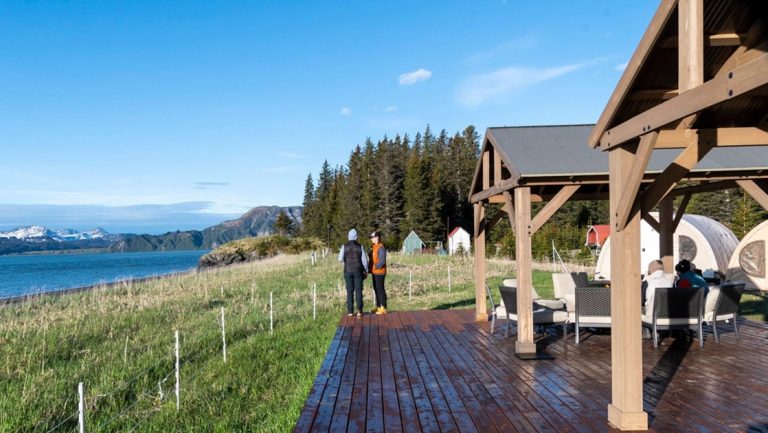 Alaska travelers stand on a wooden deck beside a gazebo, surrounded by electric fence, and gaze out onto the water on a sunny day.
