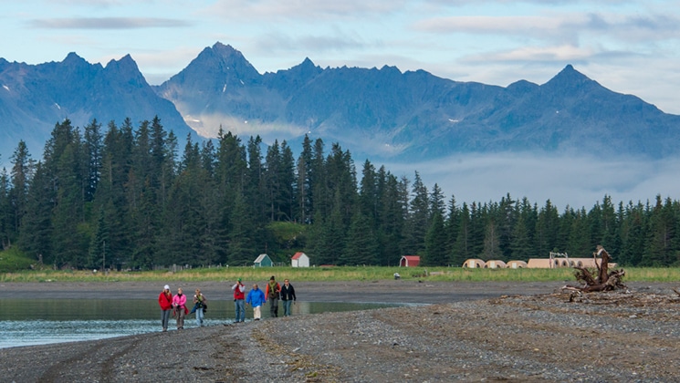 Alaska travelers walk at water's edge near to a small off-the-grid tent camp with grassland, forest & large mountains behind.