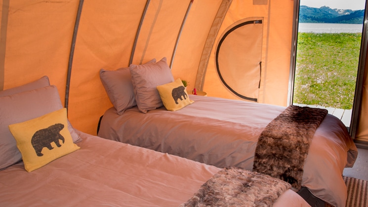 Inside of modern beige hoop tent with 2 twin beds covered in gray bedding & door open to the Alaska wilderness.