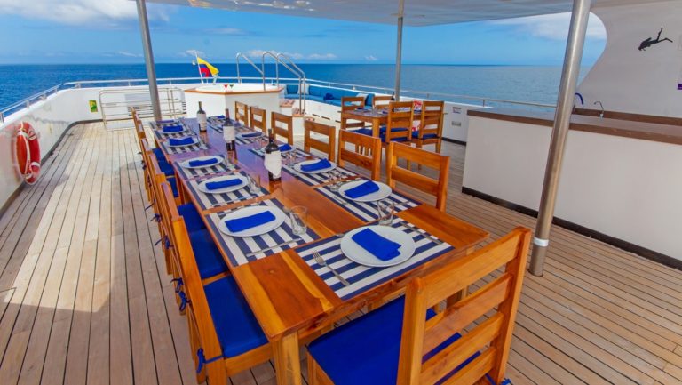 Al fresco Sundeck dining on Calipso yacht in Galapagos with teak decking & long wooden table set with blue & white linens.