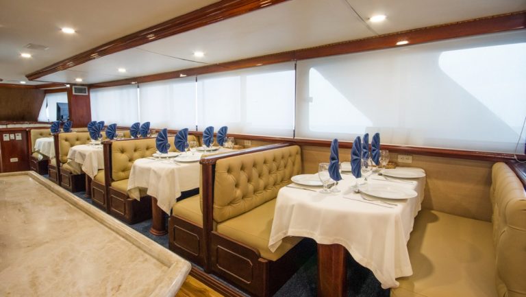 Dining room on Galapagos Sky dive boat with a row of gold-colored booths set for 4 beside a row of view windows & granite bar.