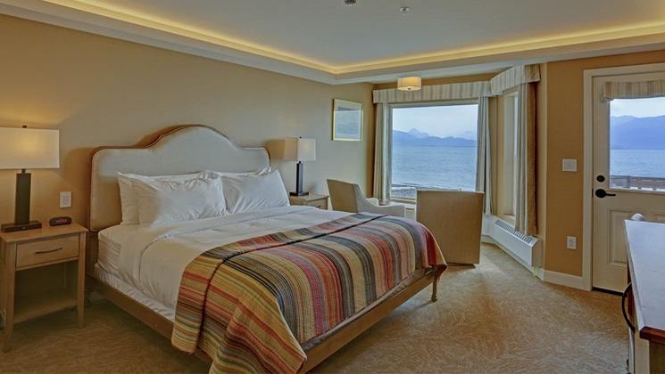 Land's End Resort guest room with king bed, sea views, beige accents, oceanfront door, plush chairs, bedside tables & wooden furniture.
