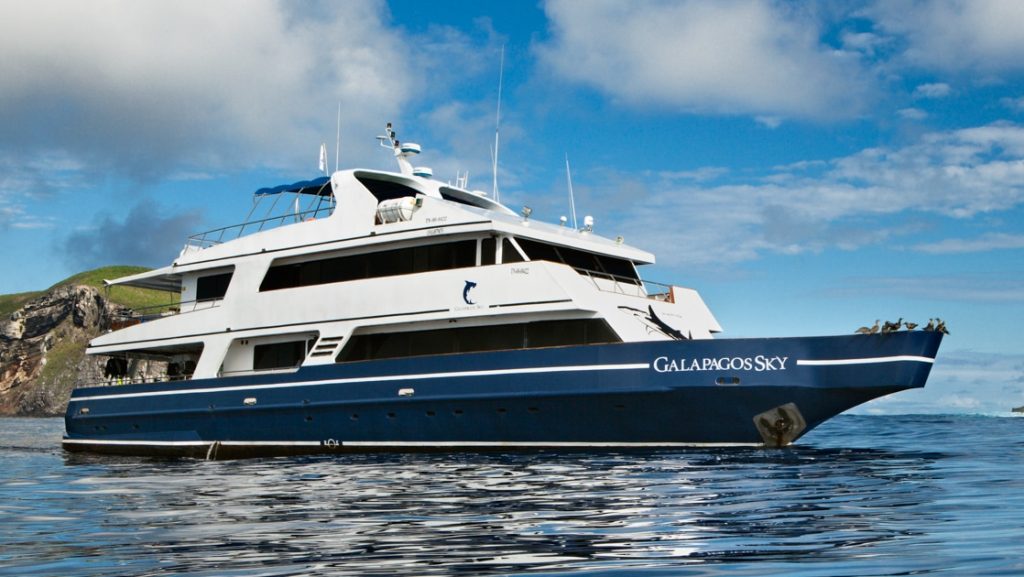 Water-level view of Galapagos Sky dive boat with blue hull & 2 white upper decks, sitting in calm water beside a green island/