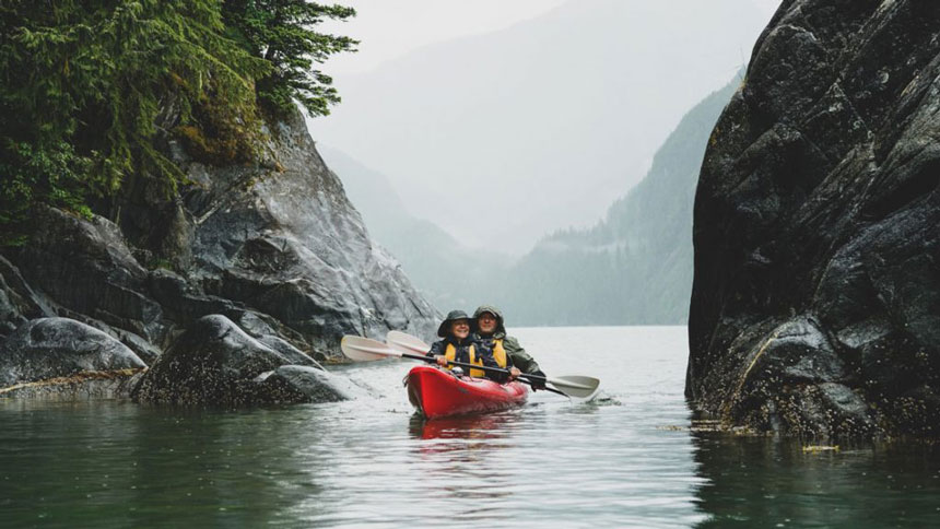 Tandem kayakers in a red boat paddle calm water between large dark boulders on a misty day during the Wild & Wooly Alaska Cruise.