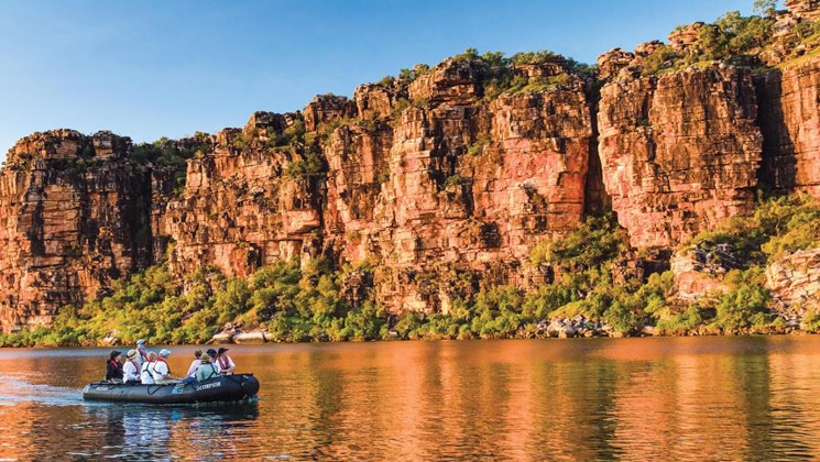 Travelers on a Nat Geo expedition cruise in a black Zodiac in glassy water beside tall red rock walls seen on Kimberley cruises.