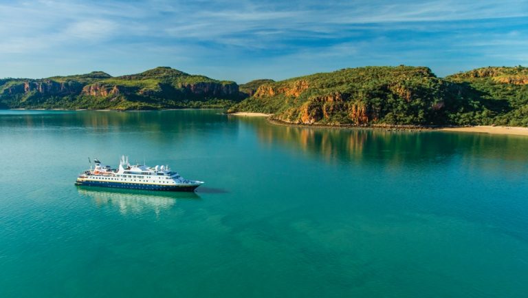 Nat Geo Orion small expedition ship with blue hull & white upper decks sits in turquoise water beside red & green islands in Australia.