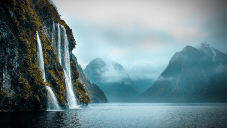 Cloudy. overcast day on the Ultimate Fiordland Cruise with calm water being fed by tall waterfalls cascading down silver rock.