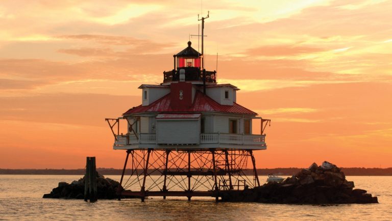 Short & stout, red & white Thomas Point Lighthouse sits on metal legs above the water under an orange sunset, seen on a Chesapeake Bay cruise.