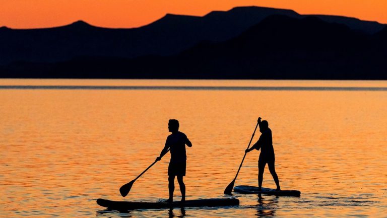 Travelers in shadow paddle stand-up paddleboards in calm water at sunset during the Exploring The Bahamas' Out Islands cruise.