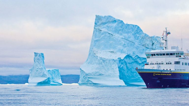 Nat Geo Explorer small ship with dark blue hull & white upper decks cruises by a large iceberg taller than the ship on the Fabled Lands of The North: Greenland to Newfoundland small ship cruise.