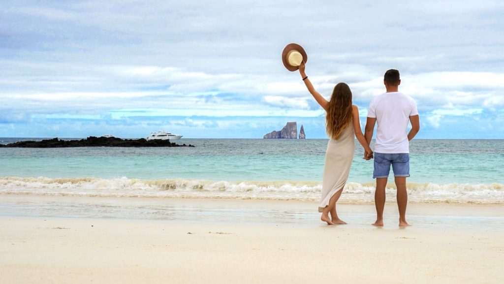 Man & woman in light-colored clothes stand on white-sand beach waving to white yacht on a Grand Majestic cruise in Galapagos.