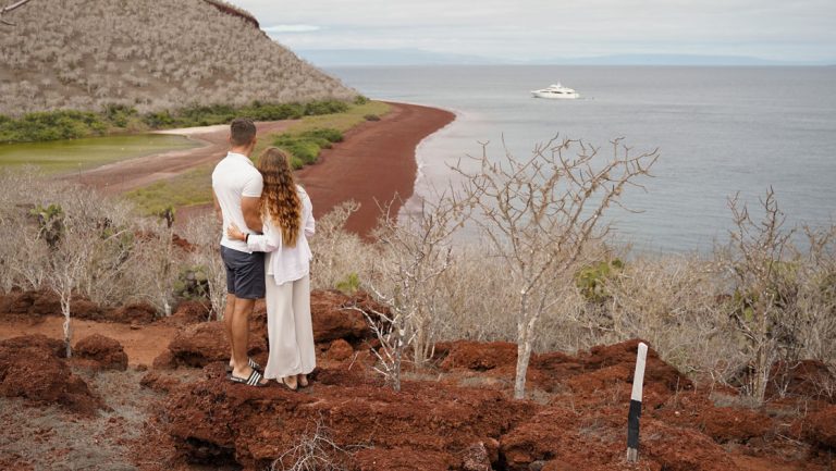 A couple stands with arms around each other overlooking the red-sand beach of Rabida Island during a Grand Majestic cruise.