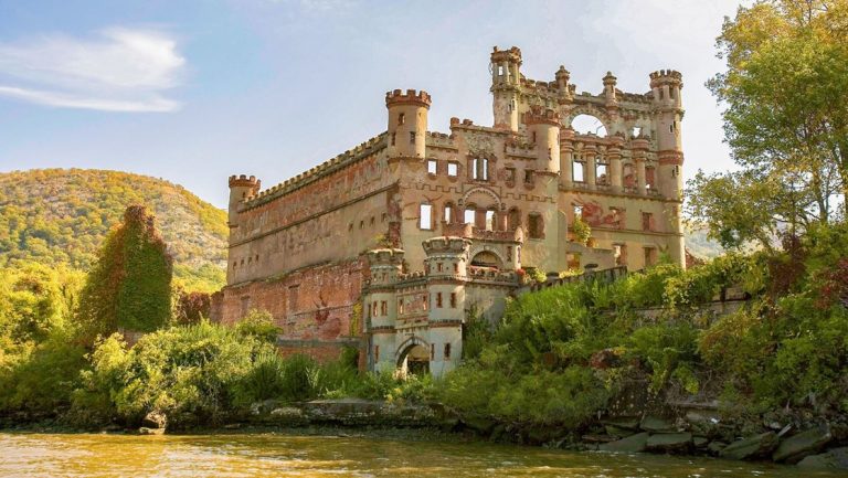 Historic castle missing half of its walls sits on a lush green riverbank at sunset, seen on a Hudson River fall foliage cruise.