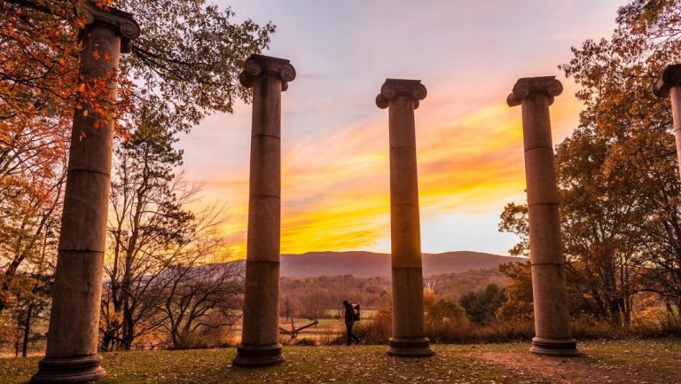 4 Roman stone pillars stand among fall foliage at sunset with mountains in the distance, seen on overnight cruises on the Hudson River.