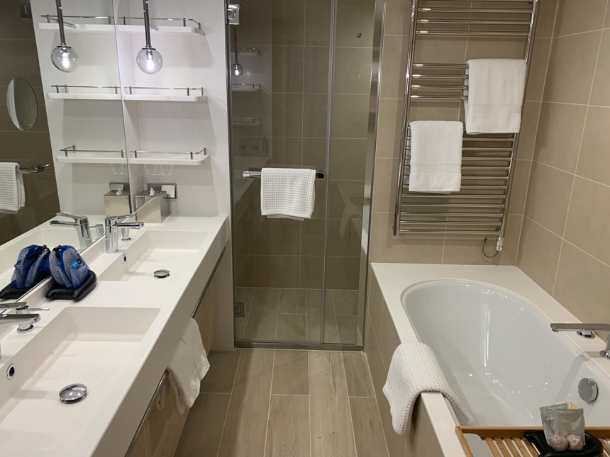 A grey White and glass cabin bathroom with white tub, separate shower, double white sinks and a silver heated towel bar.