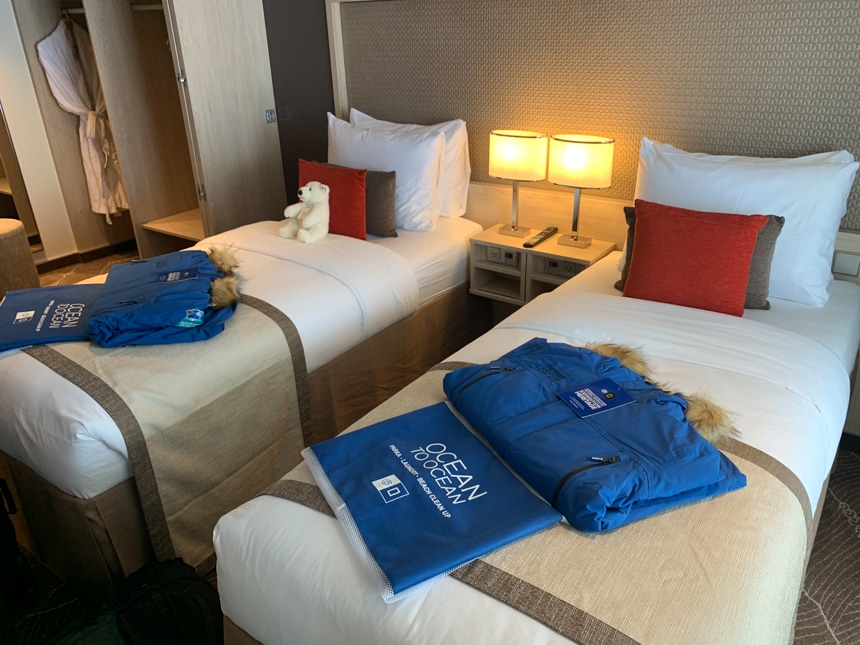 Royal blue parkas and wet bag lay on top of two double beds with white comforters inside a category 7 cabin aboard Resolution.