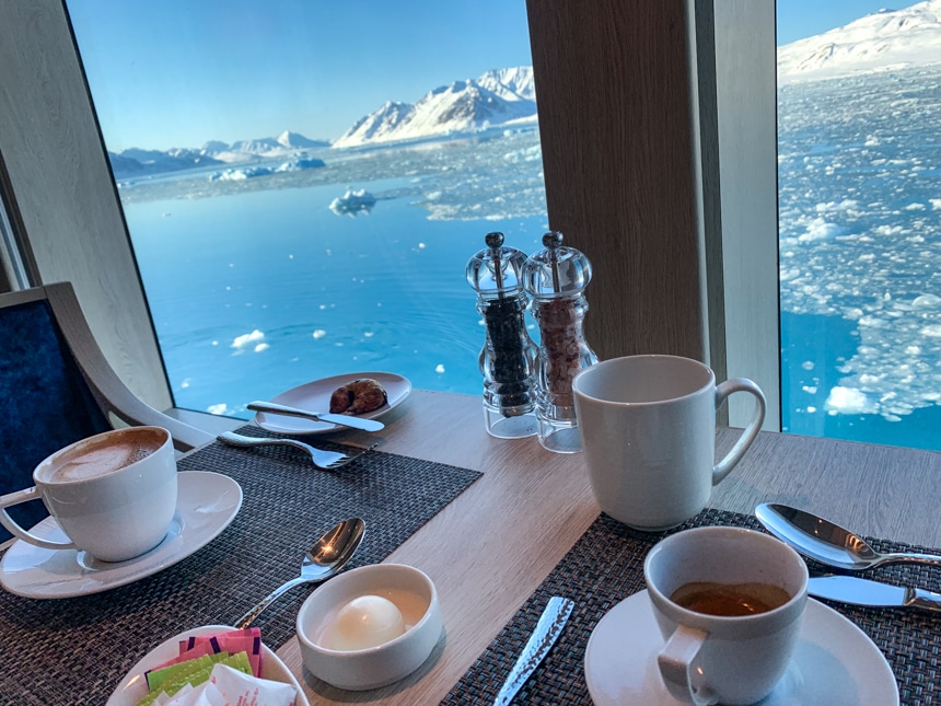 A breakfast table with cups of lattes and baked goods set against windows overlooking an arctic icy landscape on a blue sky day.