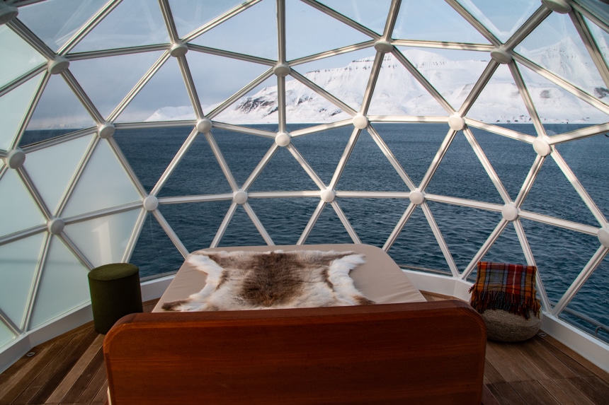 Inside the National Geographic Resolution Igloo with a bed, blanket, and ottoman, with views of an icy arctic landscape through the glass.