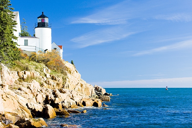 East coast cruise itineraries, near a white lighthouse on a brown rocky cliffside that leads to a blue ocean near Maine.