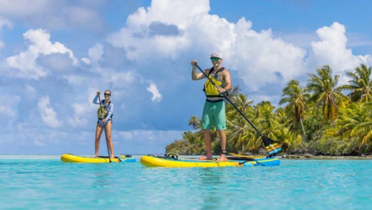 Man & woman each paddle a yellow stand-up paddleboard in turquoise water beside a small island with palm trees on a South Pacific cruise.
