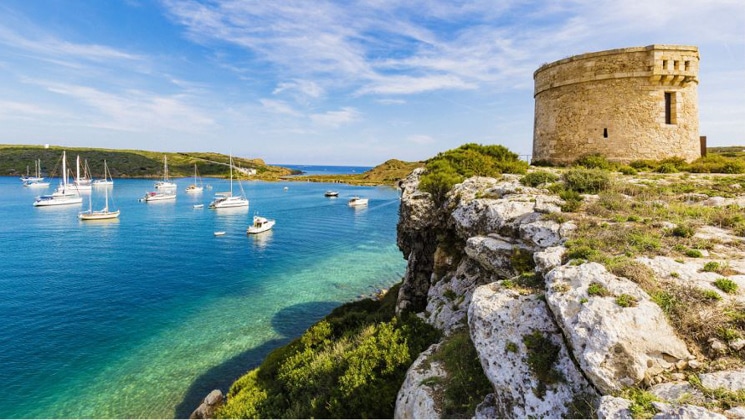Circular stone castle sits perched above a clear blue bay with white ships at anchor, seen sailing the western Mediterranean.