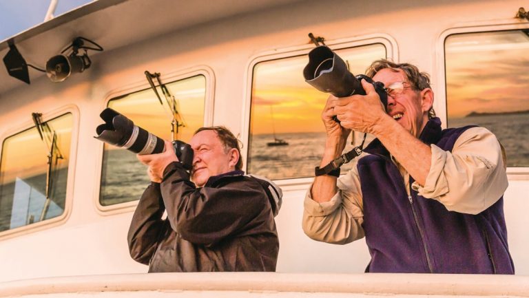2 men stand on deck of a small ship, photographing the sunset with long lenses on a Chesapeake Bay overnight cruise.