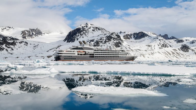 A luxury ship colored white and silver with a red stripe float in ice covered water in front of snowy mountain range in Svalbard