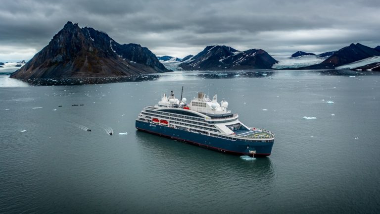 The luxury Le Commandant Charcot ship floats in a bay filled with three glacier wedged between dark rocky mountain ranges.