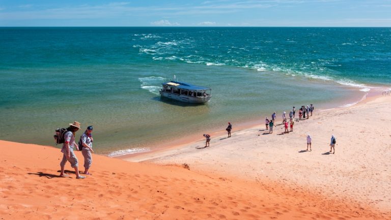 Travelers walk down a red sand hill towards people on the beach as a small aluminum boat pulls into shore in west Australia.