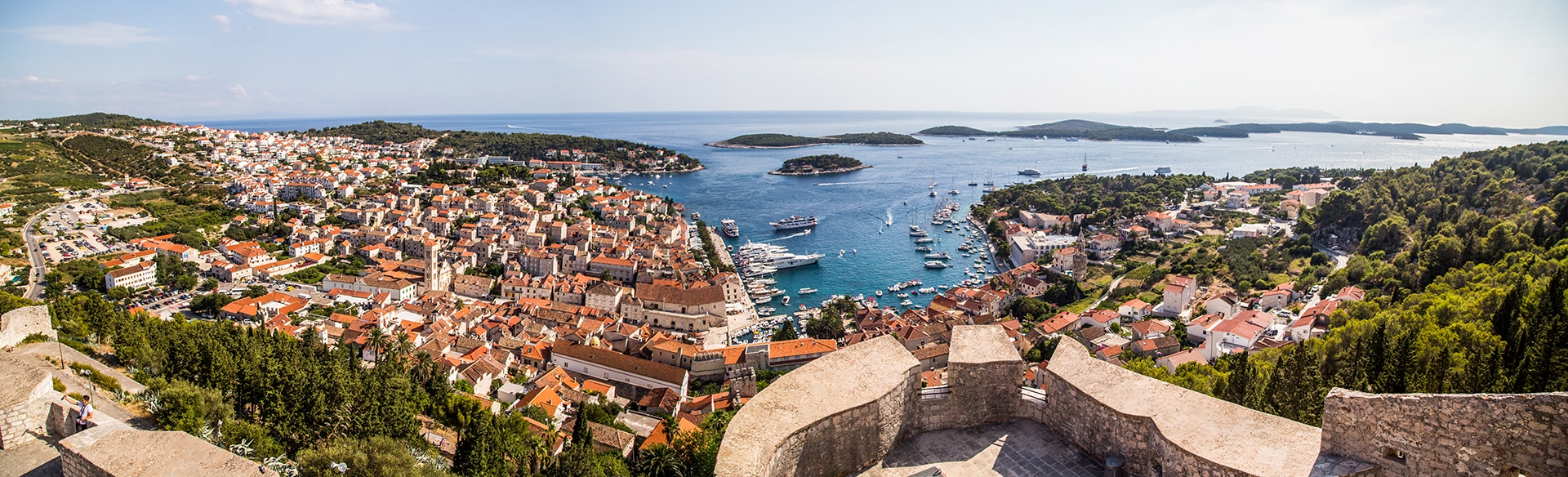Aerial view of Hvar, Croatia, showing a stone overlook, red roofs far below and small cruise ships, yachts and islets in the harbor.