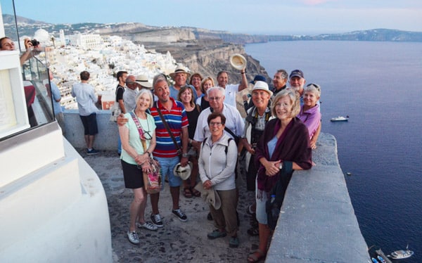 A group of small ship Mediterranean cruise travelers poses on a ledge in Santorini Greece at sunset with cliffs and white buildings behind them.